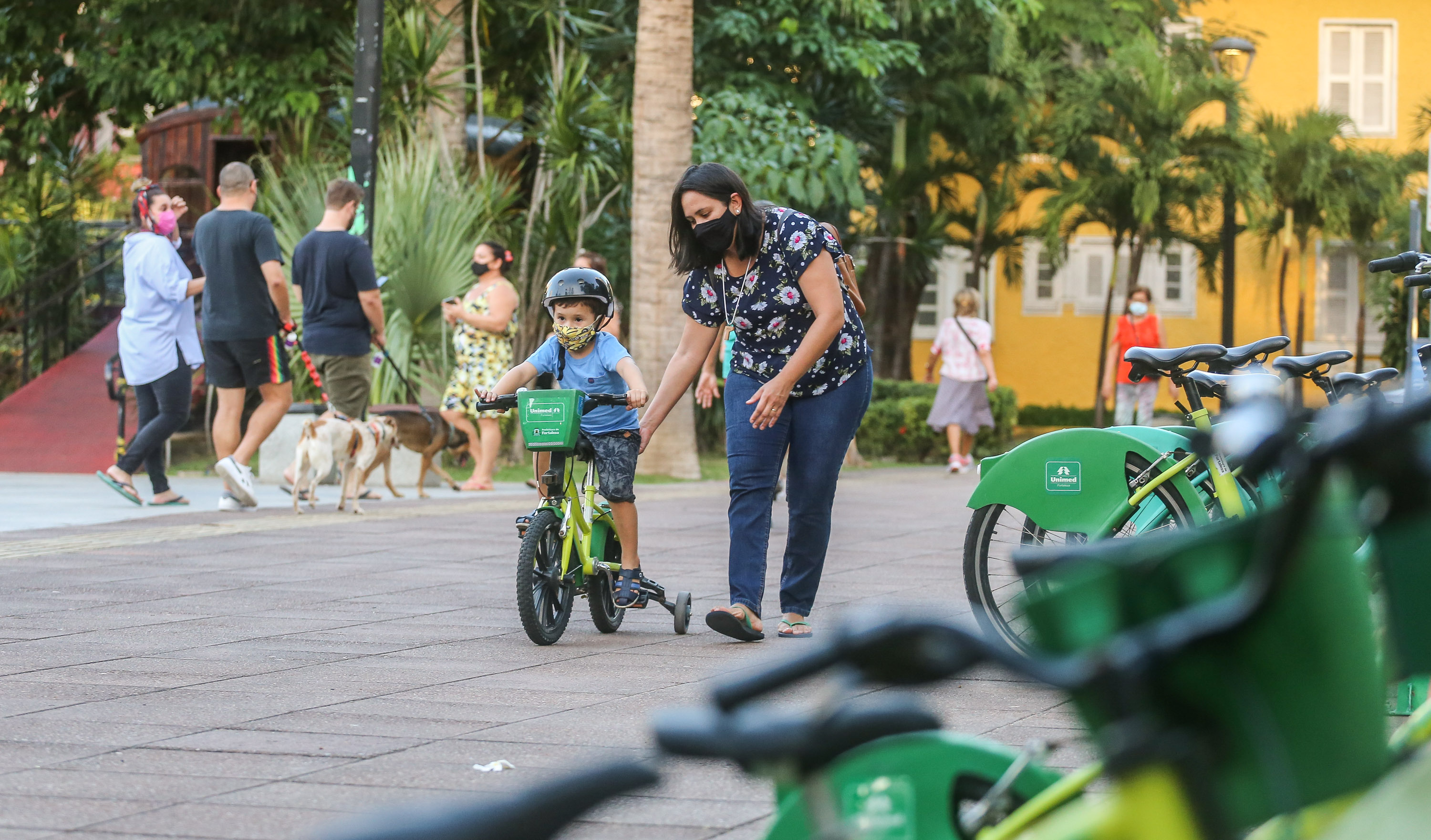 mãe acompanha o filho que anda numa bicicleta com rodinhas numa praça